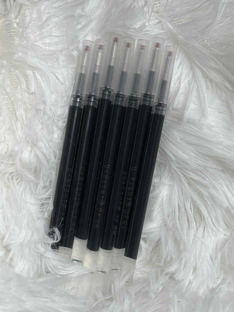 May include: A collection of black pen refills with clear plastic caps. The refills are arranged diagonally against a white, fluffy background. Each refill has a white base and a red tip.