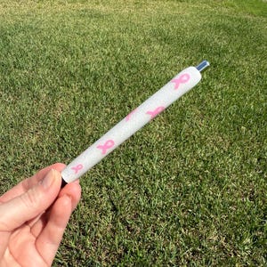 May include: A white pen with pink ribbon designs. The pen has a silver tip and a black base. The pen is held against a background of green grass.