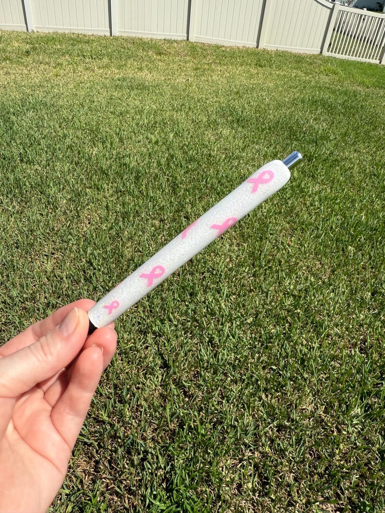 May include: A white pen with pink ribbon designs. The pen has a silver tip and a black base. The pen is held against a background of green grass.