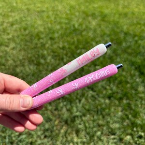Puede incluir: Se sostienen dos bol&iacute;grafos rosas con purpurina. Uno tiene el nombre "Halley" en escritura rosa sobre fondo blanco. El otro tiene la palabra "Precious" en escritura rosa. Ambos bol&iacute;grafos tienen puntas plateadas y un dise&ntilde;o de purpurina rosa.