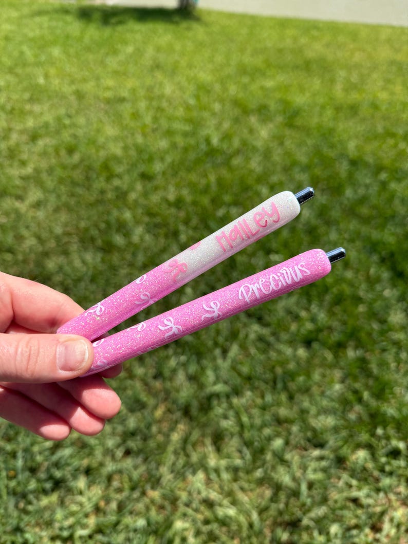 Puede incluir: Se sostienen dos bol&iacute;grafos rosas con purpurina. Uno tiene el nombre "Halley" en escritura rosa sobre fondo blanco. El otro tiene la palabra "Precious" en escritura rosa. Ambos bol&iacute;grafos tienen puntas plateadas y un dise&ntilde;o de purpurina rosa.