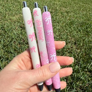 Puede incluir: Tres bol&iacute;grafos con purpurina y dise&ntilde;os de lazos rosas. Un bol&iacute;grafo es blanco, otro es rosa y otro es blanco con el nombre "Jennifer" en rosa. Los bol&iacute;grafos tienen puntas plateadas y puntas de tinta negra.