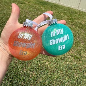 Könnte beinhalten: Zwei glitzernde Weihnachtskugeln. Eine ist orange mit dem Text "In My Showgirl Era" in Silber. Die andere ist türkis mit dem gleichen Text in Weiß. Beide haben silberne Kappen und weiße Bänder.