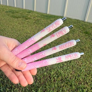 May include: Four pink and white glitter pens with silver tips are held in a hand. Each pen has a name printed on it: Skylar, Emily, Addison, and Amelia. The pens have a pink ribbon design.