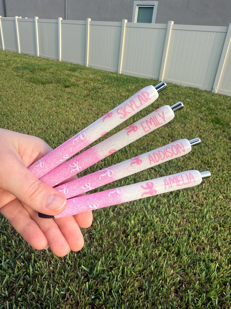 May include: Four pink and white glitter pens with silver tips are held in a hand. Each pen has a name printed on it: Skylar, Emily, Addison, and Amelia. The pens have a pink ribbon design.