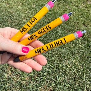 May include: Three yellow glitter pens with black text that reads "MS. LISA", "MRS. YODIS", and "MRS. LILHOLT". The pens have a silver tip, a pink band, and a black tip. The pens are held against a background of green grass.