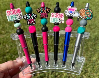 May include: Six colorful pens with different charms on top. The charms include a scrub top, a stethoscope, a heart, and a nurse's cap. The pens are displayed in a clear plastic holder.