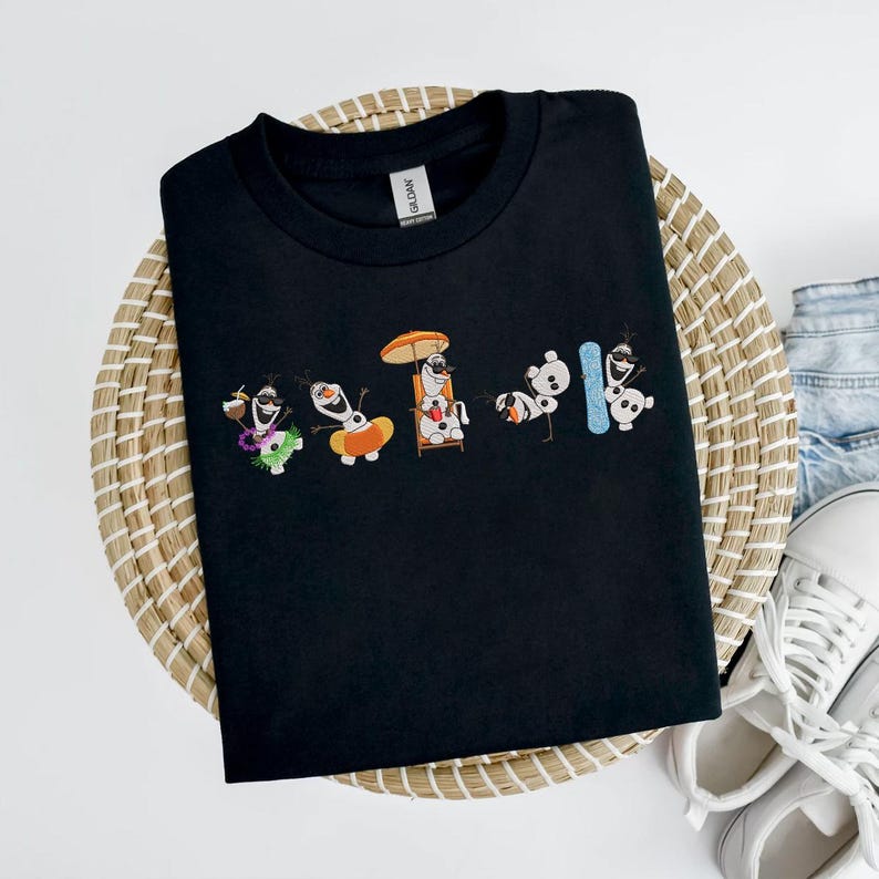May include: Black crew neck t-shirt featuring an embroidered design of Olaf from Frozen in various summer activities. Olaf is shown with a drink, on a float, in a beach chair, and with a snowboard. The shirt is displayed on a woven basket.