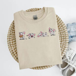 May include: Beige crew neck t-shirt with an embroidered design of four cartoon pigs in various poses. The pigs are white with black spots and pink ears. The shirt is folded on a woven basket.