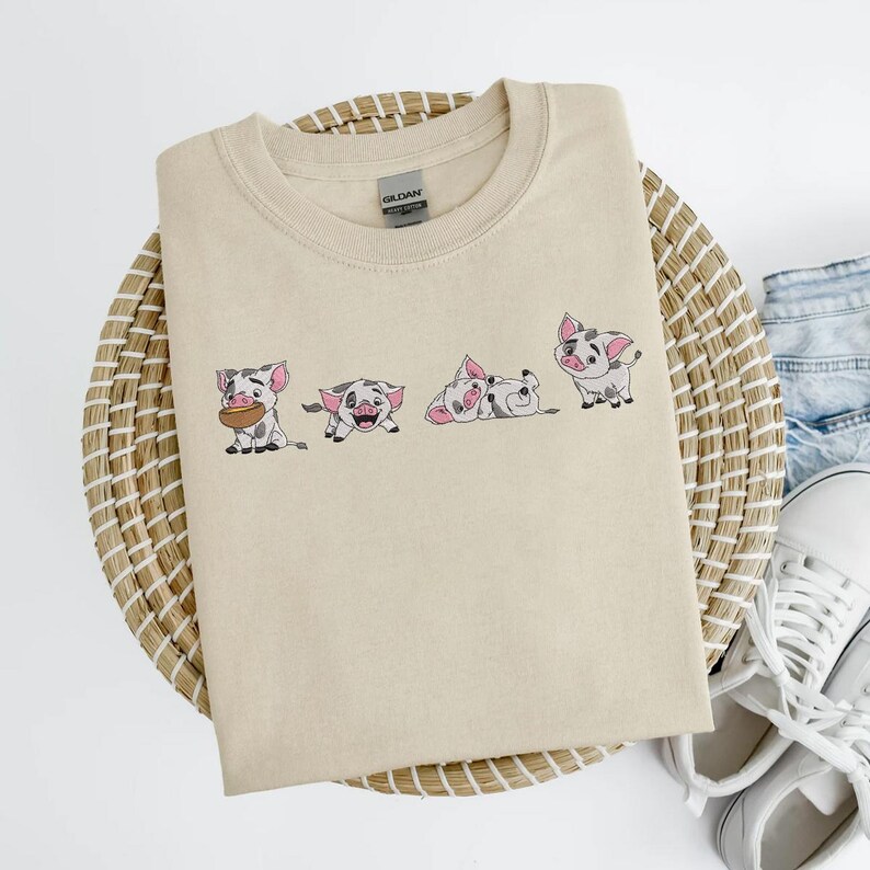 May include: Beige crew neck t-shirt with an embroidered design of four cartoon pigs in various poses. The pigs are white with black spots and pink ears. The shirt is folded on a woven basket.