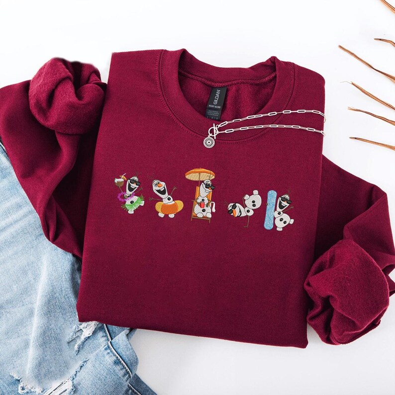 May include: A maroon crewneck sweatshirt featuring embroidered cartoon characters. The design includes several Olaf characters in various summer activities. A silver necklace with a pendant is draped on the sweatshirt.