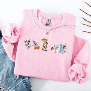May include: A light pink sweatshirt featuring an embroidered design of several Olaf characters in various summer-themed poses. The design includes Olaf with a drink, a float, a lifeguard chair, and a snowboard. A silver necklace with a sun pendant is also visible.