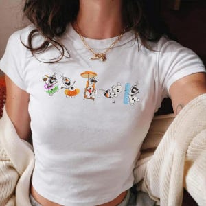 May include: White t-shirt featuring a colorful embroidered design of Olaf from Frozen enjoying various summer activities. The design includes Olaf with sunglasses, a lei, a floatie, a beach chair, and a snowboard. The shirt has short sleeves and a crew neck.