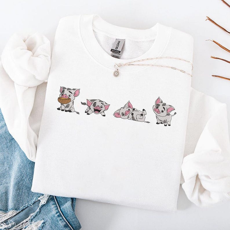 May include: White sweatshirt with four cartoon pig illustrations in various poses. The pigs are grey and pink. A silver necklace with a small pendant is also visible. The sweatshirt is folded on a white surface. The sweatshirt is made of cotton.