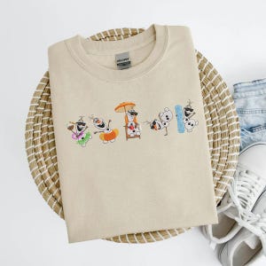 May include: Beige crew neck sweatshirt featuring an embroidered design of Olaf from Frozen in various beach-themed poses. Olaf is wearing sunglasses, a Hawaiian lei, and holding a drink. The sweatshirt is folded on a woven mat.