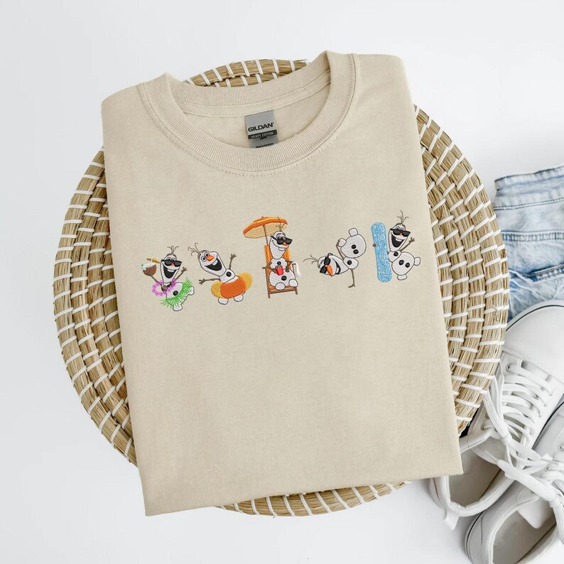 May include: Beige crew neck sweatshirt featuring an embroidered design of Olaf from Frozen in various beach-themed poses. Olaf is wearing sunglasses, a Hawaiian lei, and holding a drink. The sweatshirt is folded on a woven mat.