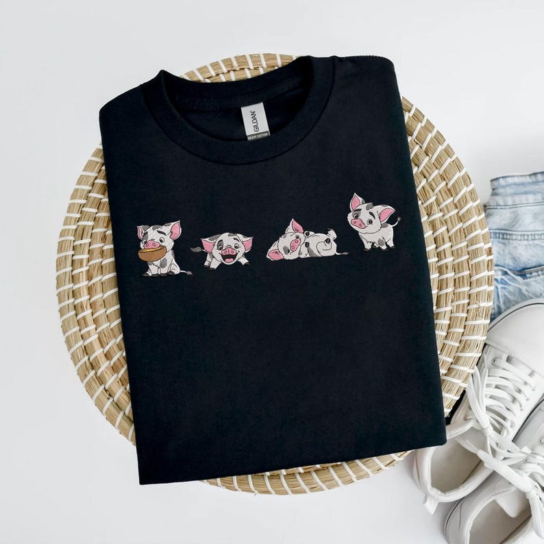 May include: Black crew neck t-shirt with four cartoon pig illustrations. The pigs are white with pink markings and in various poses. The t-shirt is displayed on a woven basket. The brand name 'Gildan' is visible.