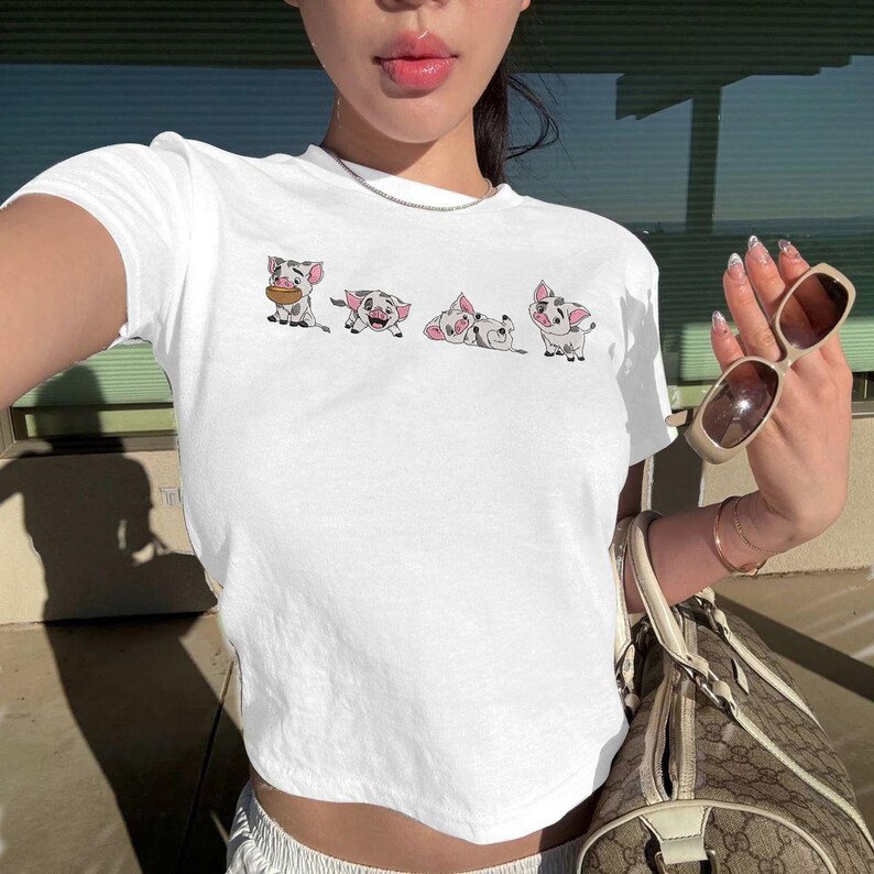 May include: White crop top with a cartoon illustration of four pigs. The pigs have pink ears and spots. The shirt is short-sleeved and has a crew neck. The top is a casual style.