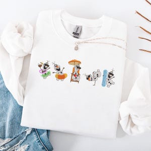 May include: White sweatshirt featuring an embroidered design of Olaf from Frozen in various summer activities. Olaf is depicted with sunglasses, a Hawaiian lei, a beach chair, and a snowboard. A silver necklace with a small pendant is also visible.