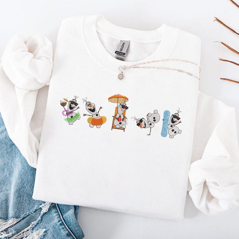 May include: White sweatshirt featuring an embroidered design of Olaf from Frozen in various summer activities. Olaf is depicted with sunglasses, a Hawaiian lei, a beach chair, and a snowboard. A silver necklace with a small pendant is also visible.