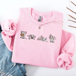 May include: A pale pink sweatshirt with an embroidered design of four cartoon pigs in different poses. The pigs are grey and white with black details. A silver necklace with a sun pendant is on the sweatshirt. The sweatshirt is folded on blue denim.