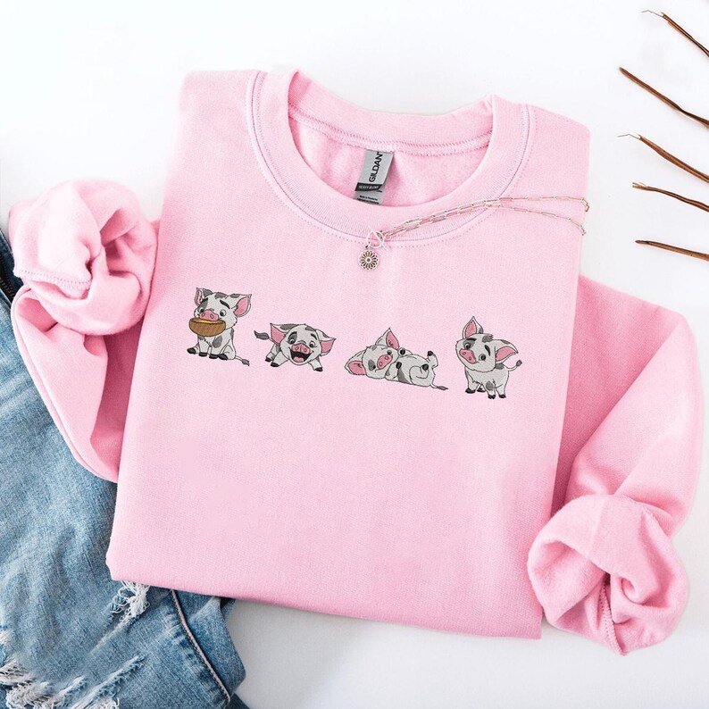 May include: A pale pink sweatshirt with an embroidered design of four cartoon pigs in different poses. The pigs are grey and white with black details. A silver necklace with a sun pendant is on the sweatshirt. The sweatshirt is folded on blue denim.
