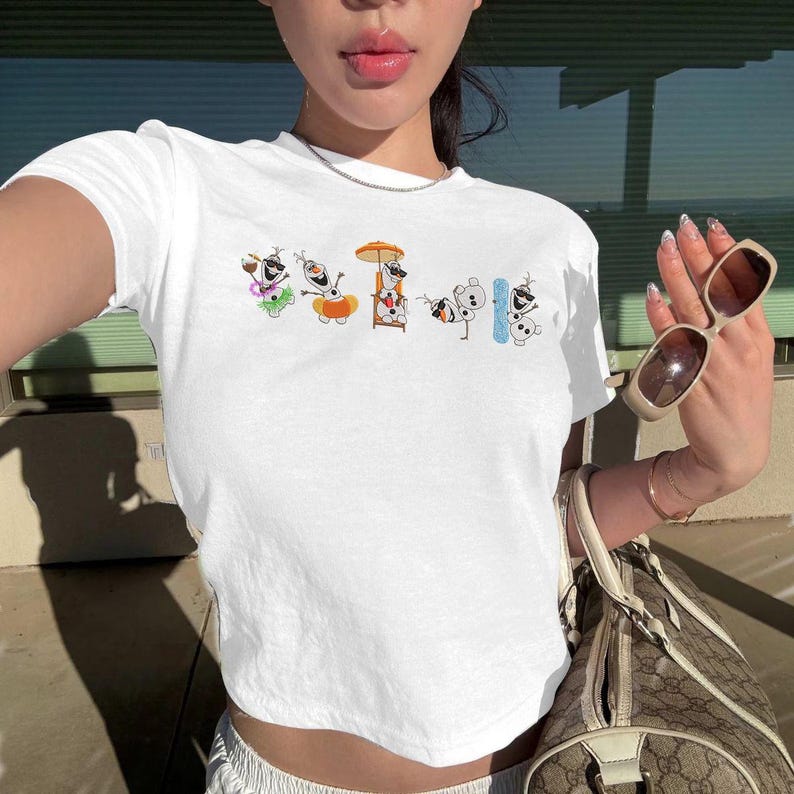 May include: White crop top featuring a colorful cartoon design of snowmen in various summer activities. The design includes snowmen with sunglasses, beach umbrellas, and snowboards. The shirt is made of a soft, comfortable material.