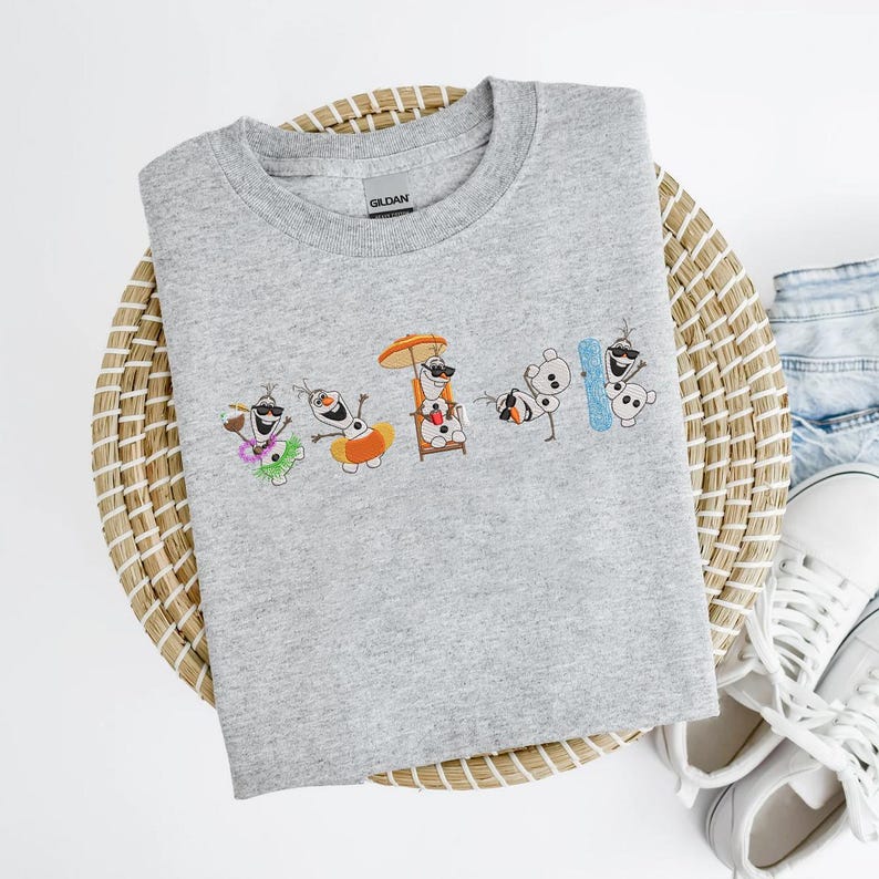 May include: A light gray crew neck sweatshirt featuring an embroidered design of Olaf from Frozen enjoying a summer vacation. The design includes Olaf in various beach-themed scenarios, such as wearing sunglasses and a Hawaiian shirt.