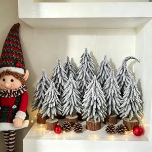 May include: A plush elf doll with a tall green and red knit hat, a plaid dress, and white trim, standing beside a collection of eleven miniature artificial pine trees with flocked white snow on their branches and round wooden bases.