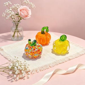Puede incluir: Tres calabazas decorativas de vidrio en diseños naranja, amarillo y multicolor. Cada calabaza tiene un tallo verde. Las calabazas están dispuestas sobre un paño color crema, con un jarrón de flores y una cinta en el fondo.