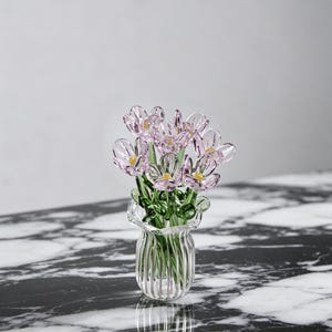Könnte beinhalten: Eine klare Glasvase mit gerüschtem Rand hält einen Strauß zarter Glasblumen. Die Blumen sind hellrosa mit gelben Zentren und grünen Stielen. Die Vase steht auf einer schwarz-weißen Marmoroberfläche.