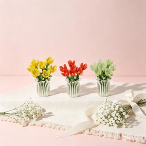 Könnte beinhalten: Drei kleine Glasvasen, jede mit einem andersfarbigen Blumenarrangement: gelb, rot und grün. Die Vasen stehen auf einem weißen Tuch, vor einem rosa Hintergrund. Ein Strauß weißer Blumen und ein Band sind ebenfalls vorhanden.