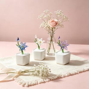 May include: A still-life arrangement featuring glass flower sculptures in small white square pots. A pink rose and baby's breath are in a clear vase. The flowers are blue, white, and purple. The background is a soft pink.