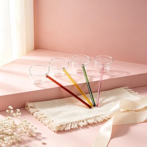 Könnte beinhalten: Vier klare Glasschalen mit kleinen Ausgießern, jede mit einem langen, schlanken Griff in einer anderen Farbe: Dunkelrot, Gelb, Grün und Hellrosa. Die Schalen sind in einer Reihe angeordnet und zeigen ihr transparentes, abgerundetes Design.