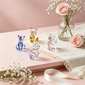 Puede incluir: Una colección de pequeñas figuras de gato de cristal de colores. Los gatos son de varios colores, incluyendo azul, ámbar, rosa y morado. Las figuras están dispuestas sobre una superficie rosa con un paño blanco y un jarrón de rosas rosas.