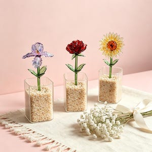 May include: Three glass flower sculptures: an iris, a rose, and a sunflower, each in a square glass vase filled with sand. The iris is purple, the rose is red, and the sunflower is yellow. A small bouquet of white flowers with a ribbon is in the foreground.