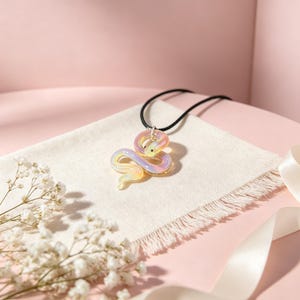 Op de afbeelding: Een ketting met een glazen slangenhanger in geel, roze en paarse tinten. De hanger hangt aan een zwart koord en ligt op een witte doek. De achtergrond is roze, met witte bloemen en een lint.