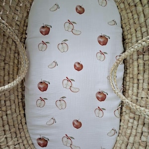 Peut inclure: Une housse de matelas en tissu blanc ornée d'un motif répétitif de pommes rouges entières et de moitiés de pommes avec des tiges vertes, placée dans un panier en osier naturel tressé avec des anses latérales.