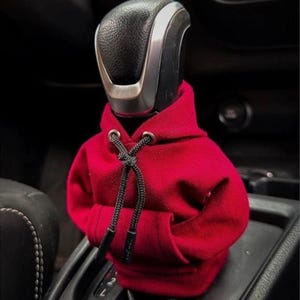 Op de afbeelding: Een rode miniatuur hoodie bedekt de versnellingspook van een auto. De hoodie heeft een trekkoord en een klein zakje. De versnellingspook is zwart en zilver. De achtergrond is donker, met het interieur van de auto zichtbaar.