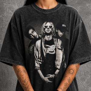 May include: A charcoal grey acid-wash t-shirt featuring a black and white graphic print of the band Nirvana. The design displays the three band members, with the central figure wearing round sunglasses and a patterned jacket over a graphic t-shirt.