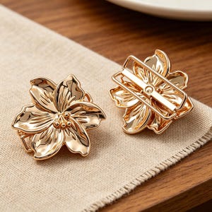 Könnte beinhalten: Zwei goldfarbene, blütenförmige Serviettenringe. Jeder Ring hat ein detailliertes Blumendesign mit Blütenblättern und einem zentralen Cluster. Die Ringe sind aus glänzendem Metall gefertigt und liegen auf einer beigen Leinen-Serviette.