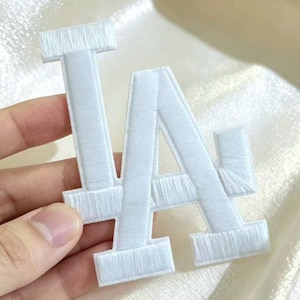 May include: A white embroidered patch featuring the overlapping letters L and A in a stylized font. The patch has a textured, stitched surface with a clean white thread finish, designed for application onto fabric surfaces as a decorative element.