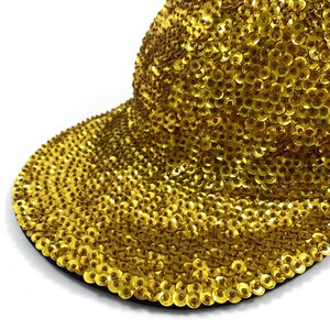 Puede incluir: Una gorra de béisbol de lentejuelas doradas. La gorra está hecha de un material suave y flexible y está cubierta de pequeñas lentejuelas brillantes.