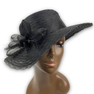 May include: A black wide-brimmed hat with a black flower and feather embellishment. The hat is made of a textured fabric and has a wide brim that is slightly angled.