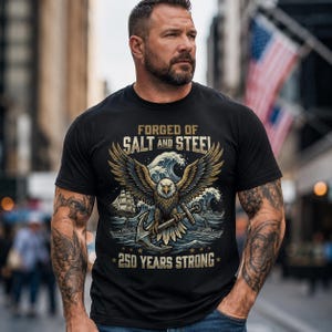 Może przedstawiać: Czarny t-shirt z grafiką przedstawiającą orła, fale i statek. Tekst głosi "Forged of Salt and Steel" i "250 Years Strong". Osoba nosząca koszulkę ma tatuaże. Tło przedstawia ulicę.