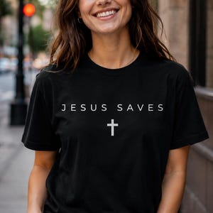 May include: Black t-shirt with the text "JESUS SAVES" and a white cross. The shirt is worn with blue jeans. The person is smiling.