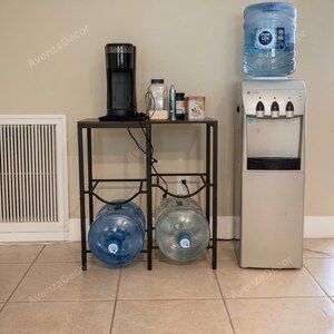 May include: A black metal stand holds a black coffee maker and various containers on top, with two large clear plastic water jugs stored on the bottom shelf. Beside the stand is a silver water cooler with a large blue-labeled water jug on top.