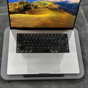 May include: A silver laptop with a black keyboard and trackpad sits on a gray carbon fiber textured lap desk. The screen displays a vibrant landscape photograph of rolling hills and fields under a bright sky. The word AvenzaDecor is visible.
