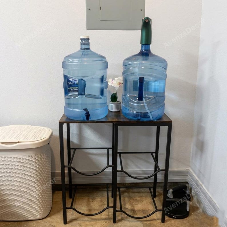 May include: A dark brown wooden water jug stand with a black metal frame featuring two tiers. Two large clear plastic water jugs sit on the top shelf, one with a white cap and one with a green electronic pump dispenser. A small white artificial flower sits between them.