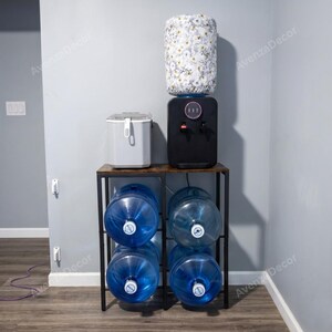 May include: A black metal water cooler stand with a wooden top shelf holding a black water cooler and a white appliance. The cooler features a floral patterned fabric cover on the water jug. Four large blue plastic water jugs are stored on the lower shelves.